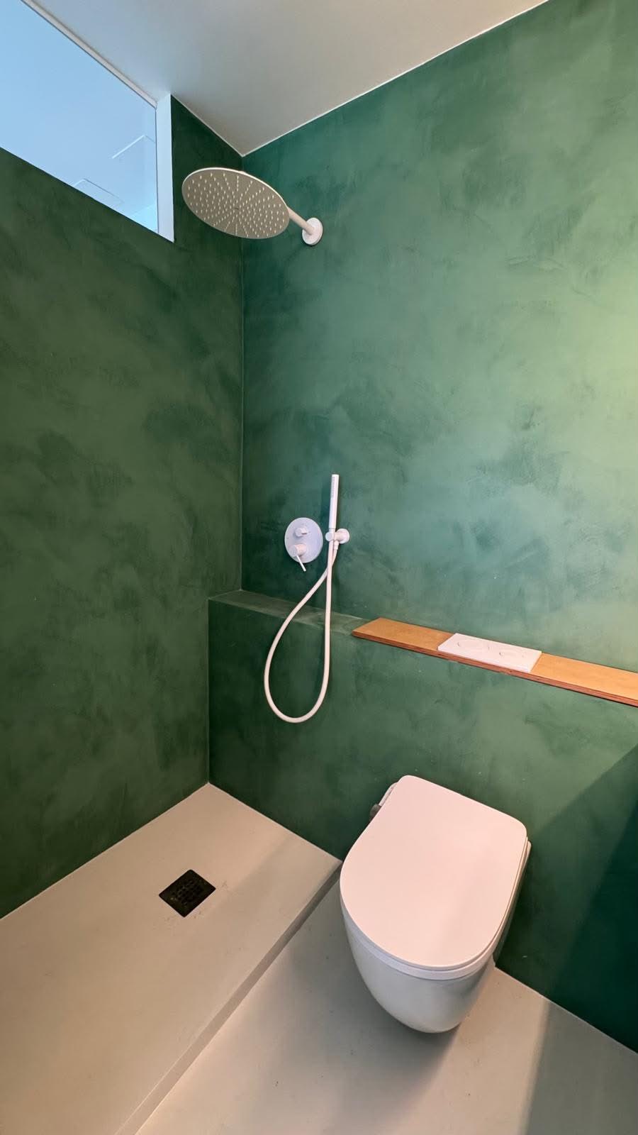 Zona de ducha con rociador de techo, grifería blanca y estante de madera, paredes en verde.
