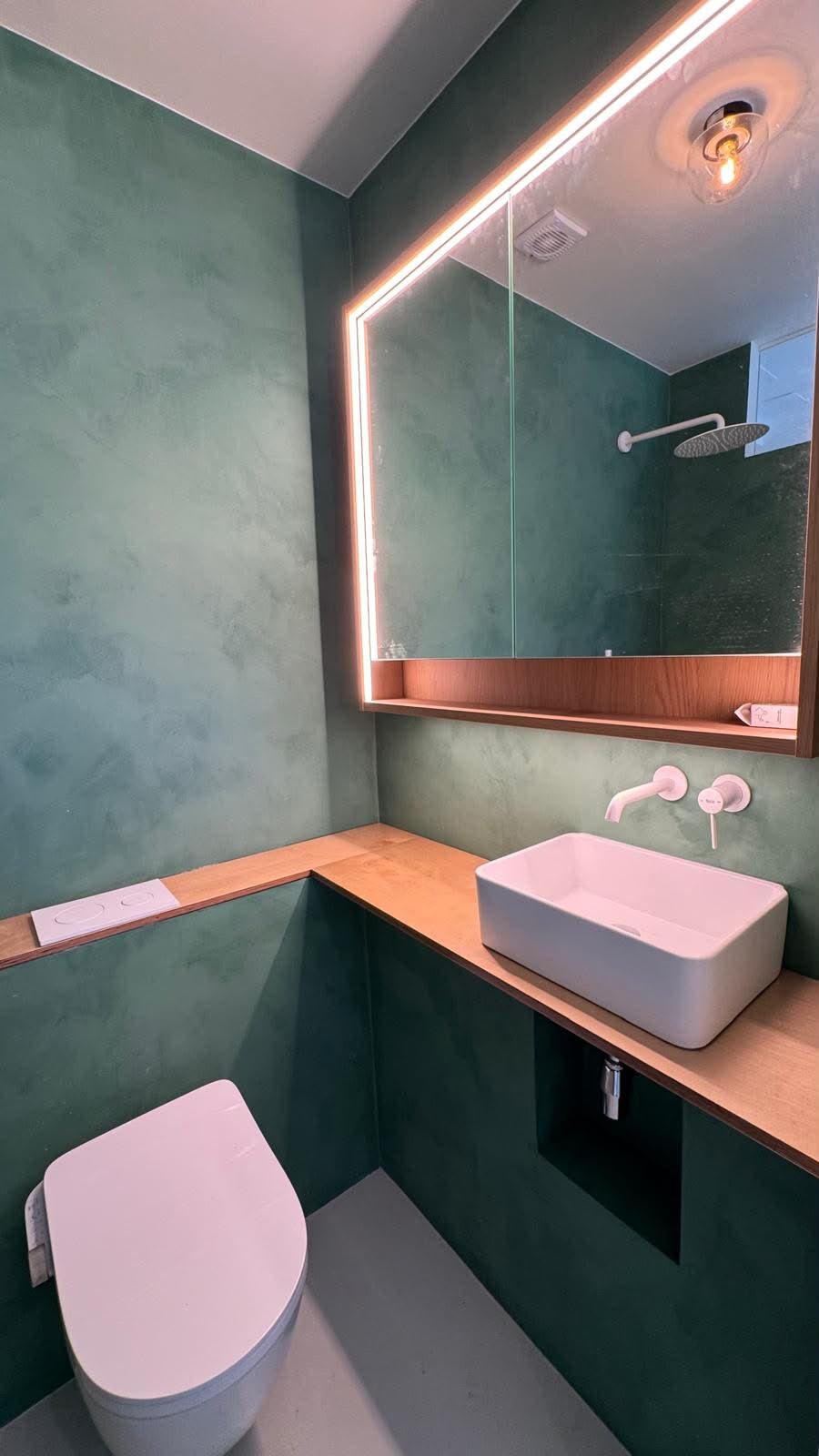 Baño con paredes verdes, espejo con luz perimetral, lavabo sobre encimera de madera e inodoro suspendido.