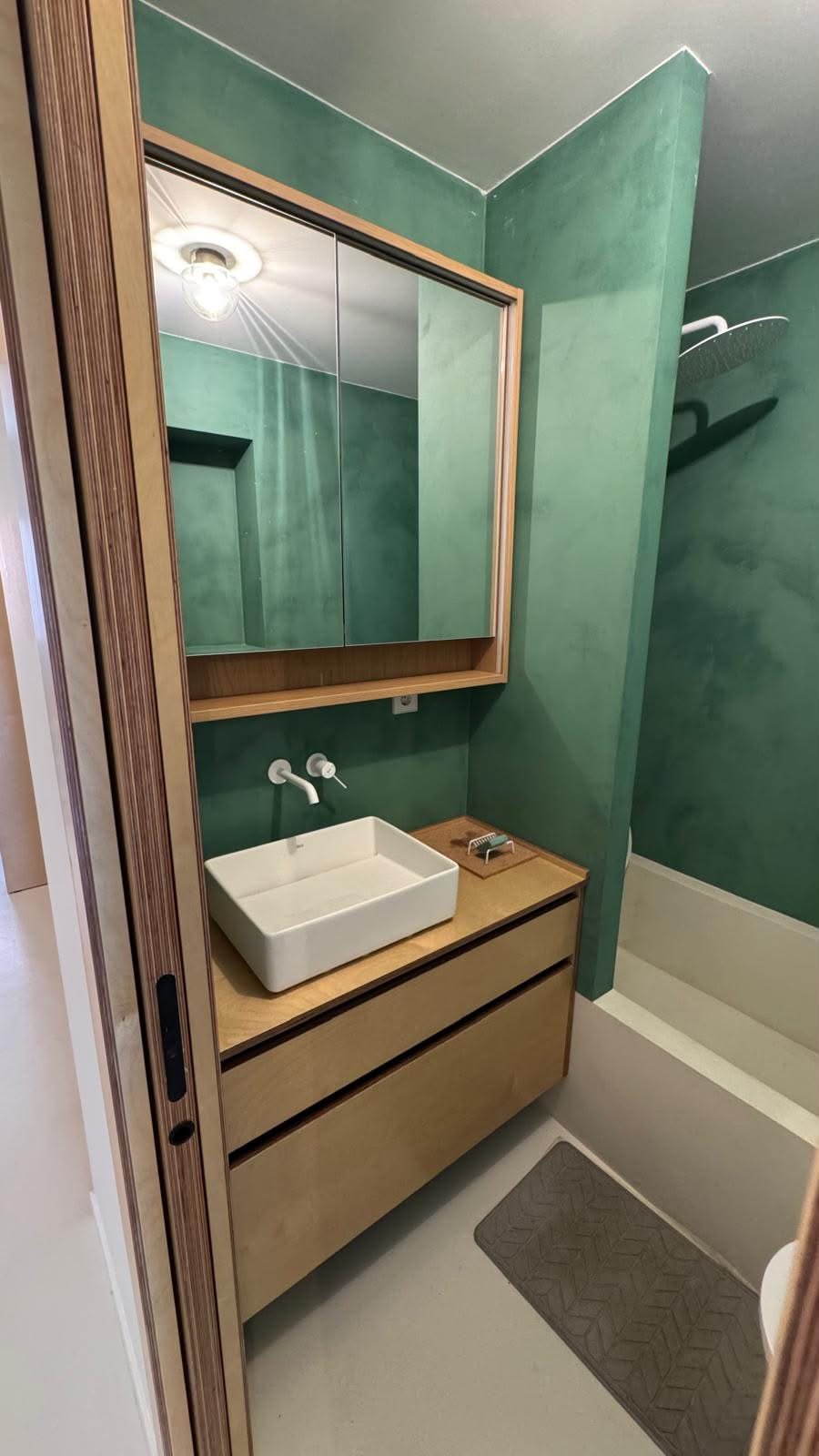 Baño con mueble de lavabo en madera, espejo de dos puertas y bañera de obra al fondo.