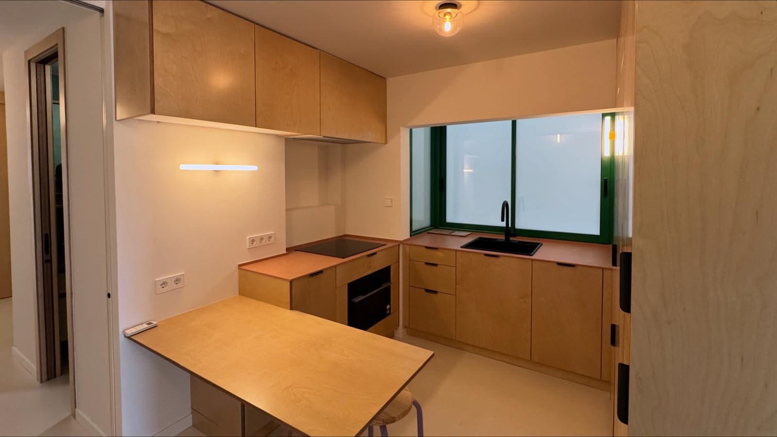 Vista general de cocina en madera con barra, fregadero negro y ventana de carpintería verde.