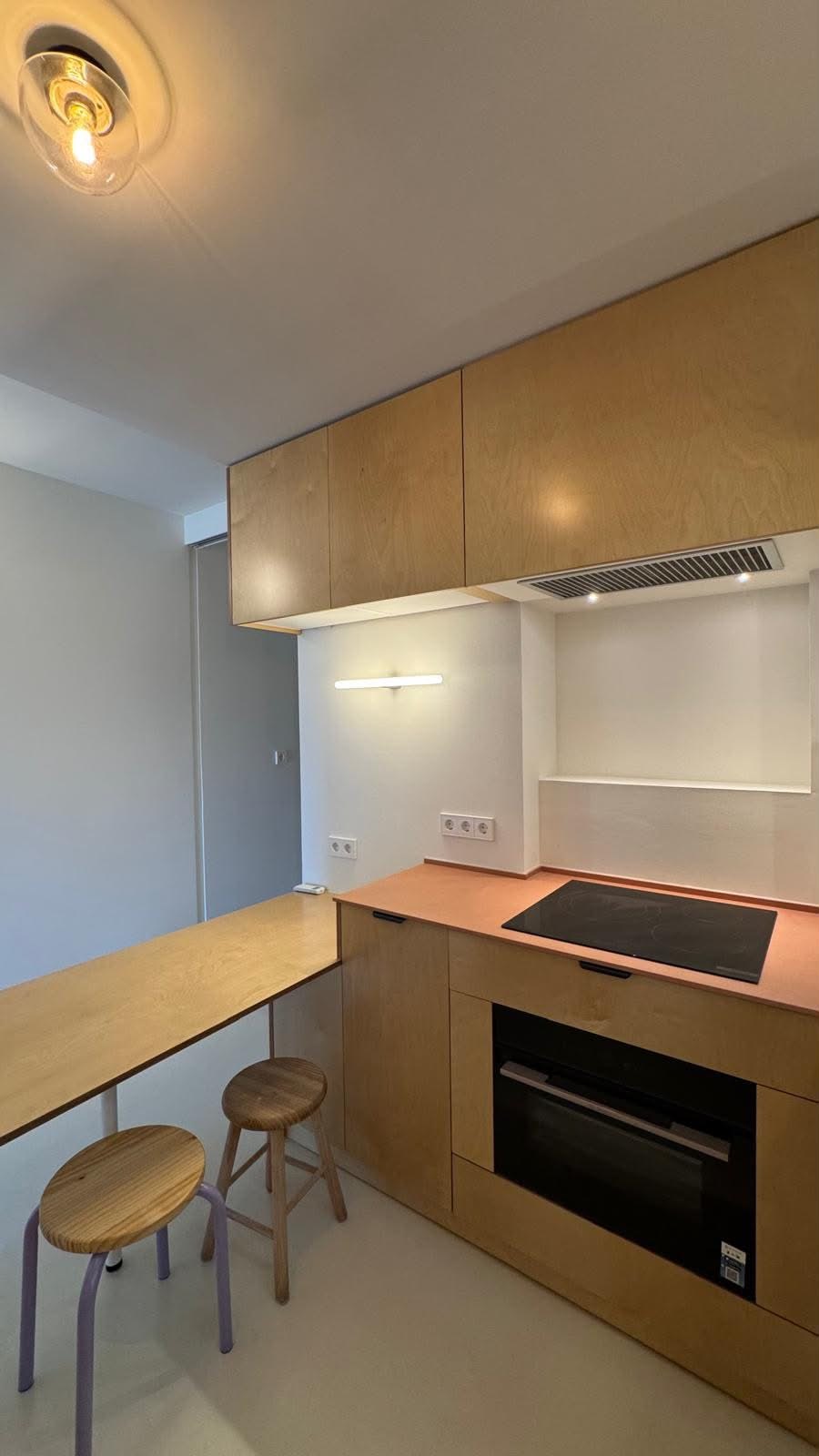 Cocina de madera con placa de inducción, horno integrado y barra con dos taburetes.